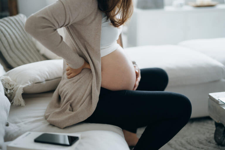 A woman sitting on the couch rubbing her belly as she thinks about her concerns regarding her high-risk pregnancy.
