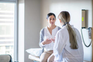 A young female of Indian ethnicity sits on the examination table at her gynaecologist's office. She is talking to her doctor about her health history.